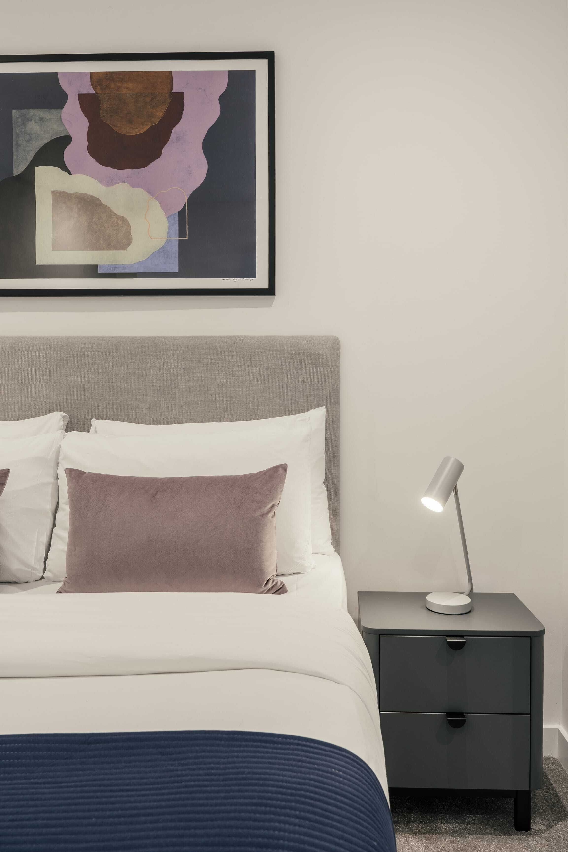 Un lit bien fait, avec des oreillers blancs et bruns, une tête de lit grise et une table de chevet sur laquelle se trouve une lampe.