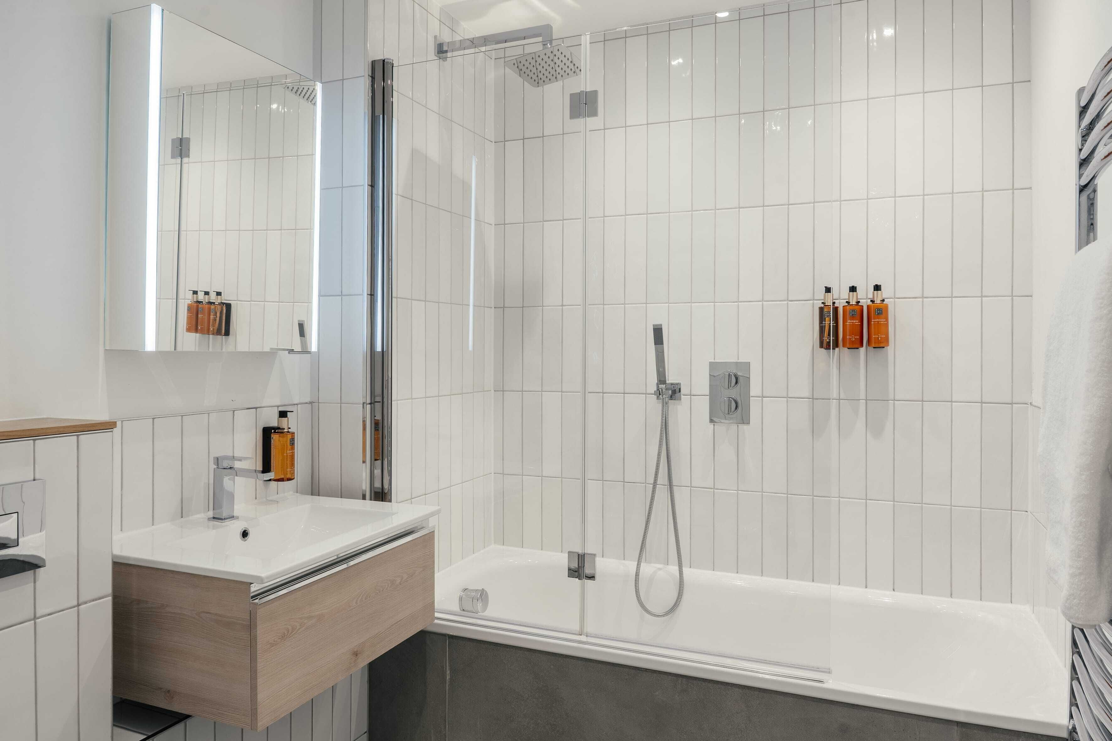 Een moderne badkamer met een witte betegelde douche, een witte badkuip en een houten wastafelmeubel met spiegel.