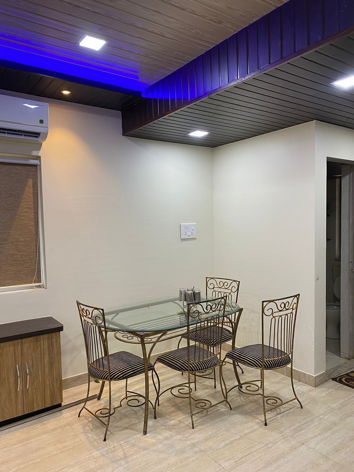 A dining area with a glass table and metal chairs, a wooden cabinet, and a ceiling with blue lighting.