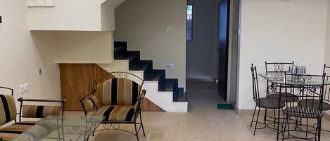 Amplia sala de estar con una mesa de centro de vidrio, sillas acolchadas, una mesa de comedor con sillas, una escalera y una puerta que da a otra habitación.