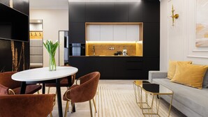 A modern kitchen with a black countertop, white cabinets, and a wooden backsplash. There is a dining area with a round table and chairs, and a sofa with yellow cushions. - Native Apartments Kołłątaja 12 (Kraków)