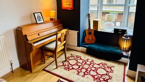 Tunnelmallinen huone, jossa on puinen piano, kitara, kehystetty kuva, punavalkoinen kuvioitu matto ja ikkuna, josta on näkymä ulos.