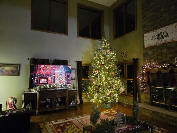 A living room with a large Christmas tree, a fireplace, and a television displaying a festive scene.