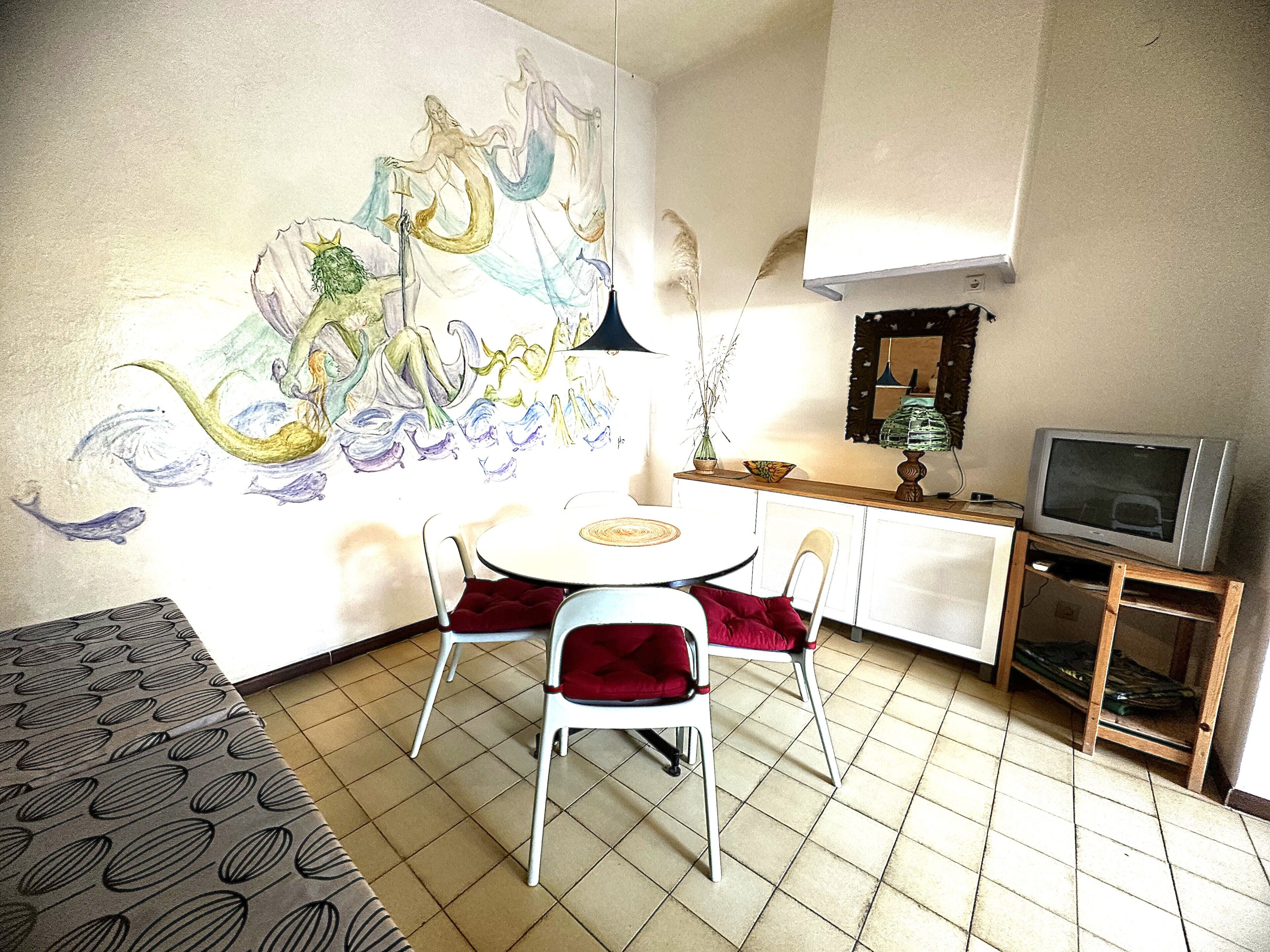 Una habitación pequeña y bien iluminada con una mesa de comedor y sillas, un televisor sobre un mueble bajo, y un gran mural abstracto en la pared.
