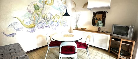Una habitación pequeña y bien iluminada con una mesa de comedor y sillas, un televisor sobre un mueble bajo, y un gran mural abstracto en la pared.
