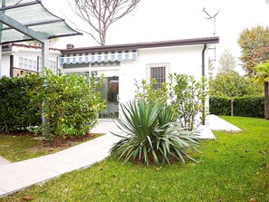 Ein modernes Haus mit weißer Fassade, einer überdachten Veranda und einem gepflegten Rasen mit verschiedenen Sträuchern und einer großen Agavenpflanze.
