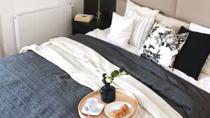 Una cama bien hecha con edredón gris y blanco, una bandeja con una taza y un pastelito, y una lámpara de noche.