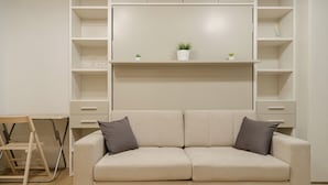 Una sala de estar minimalista con un sofá color beige, dos cojines, una planta pequeña y un escritorio plegable con una silla.