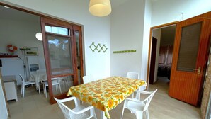 A dining area with a table covered in a floral tablecloth, surrounded by white plastic chairs. There is a wooden door and a white wall with a decorative item above it. - Holiday Villa With Private Garden Close to the sea (San Michele al Tagliamento)