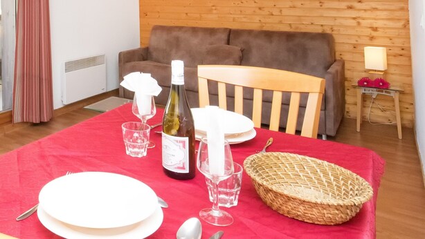 A dining area with a red tablecloth, wooden chairs, a bottle of wine, glasses, and a basket. - Apartment in Orelle Near 3 Vallees Ski Area (Orelle)