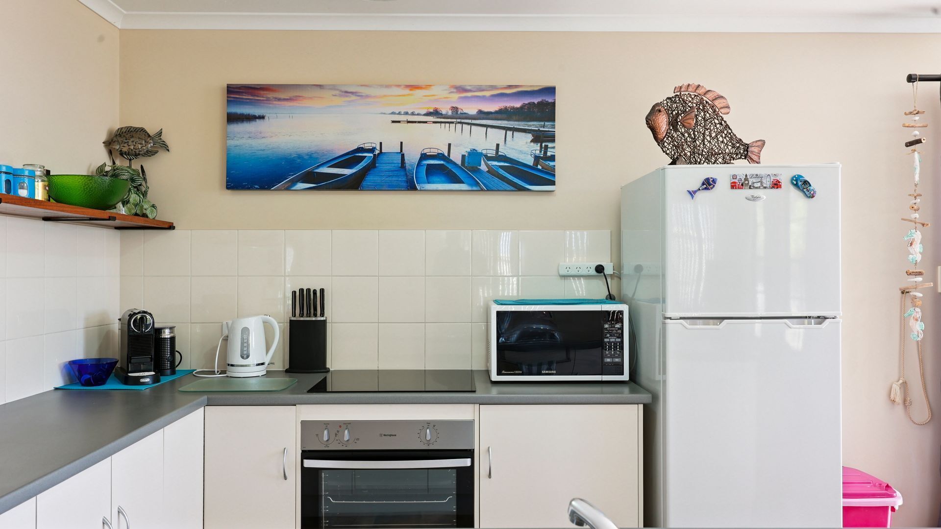 A compact kitchen with a microwave, oven, and refrigerator. There's a painting of boats on the wall and a decorative fish sculpture.