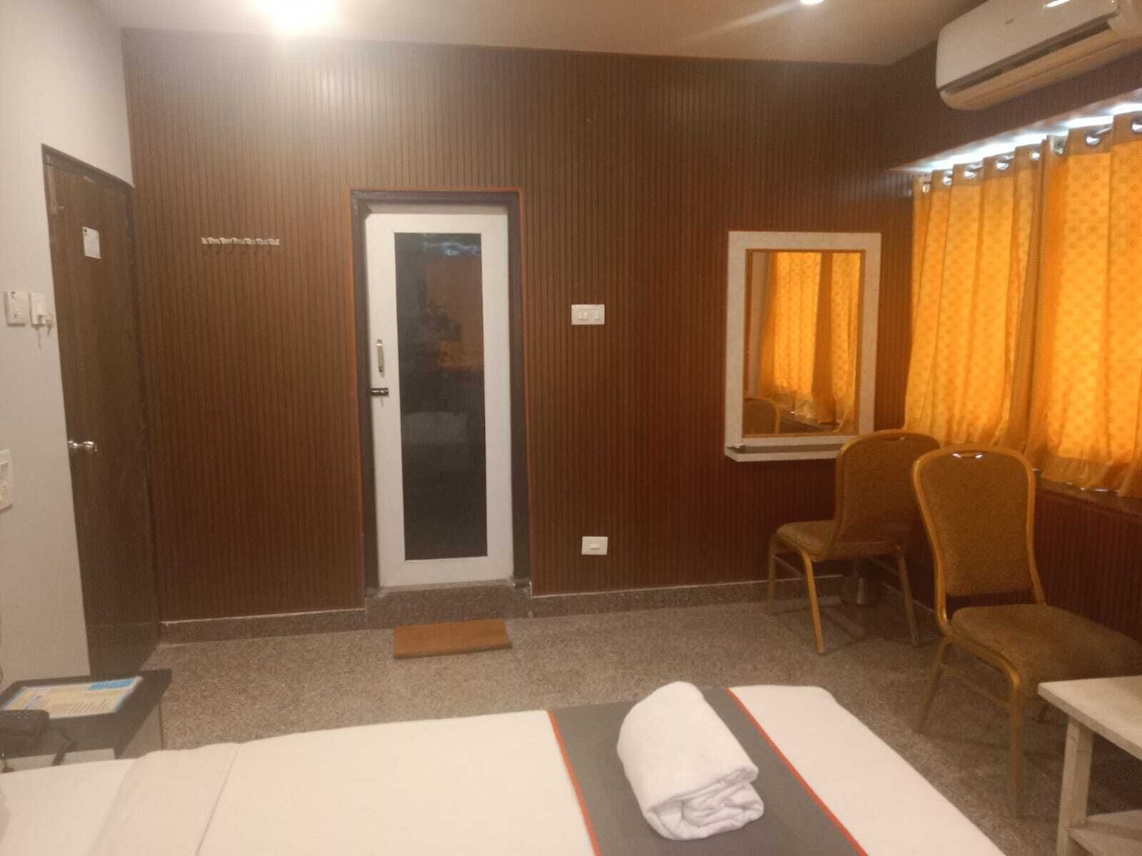 Une chambre d’hôtel avec un mur lambrissé en bois, une porte avec un panneau de verre, une fenêtre avec des rideaux jaunes, deux chaises, un lit recouvert d’une courtepointe à rayures blanches et orange, et un appareil de climatisation fixé au mur.