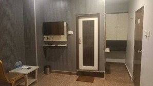 A room with a television mounted on the wall, a door, a small table with a chair, and a trash can. - Hotel Vaishnavi (Hyderabad)