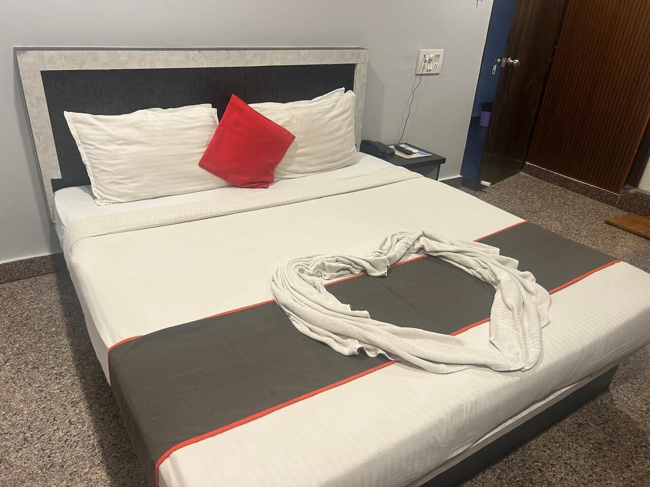 A neatly made bed with a red pillow, a white towel, and a grey and red bedspread.