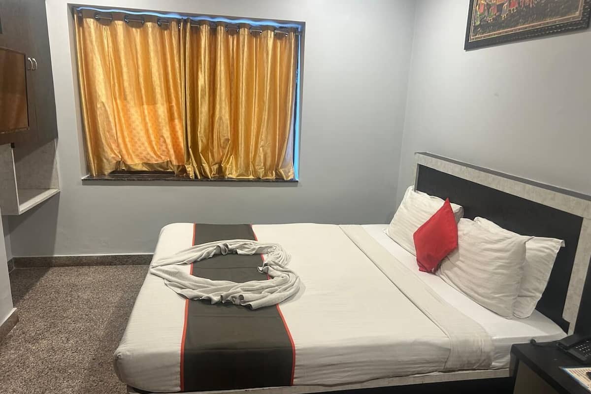 A hotel room with a bed, a nightstand, a window with curtains, and a wall-mounted painting.
