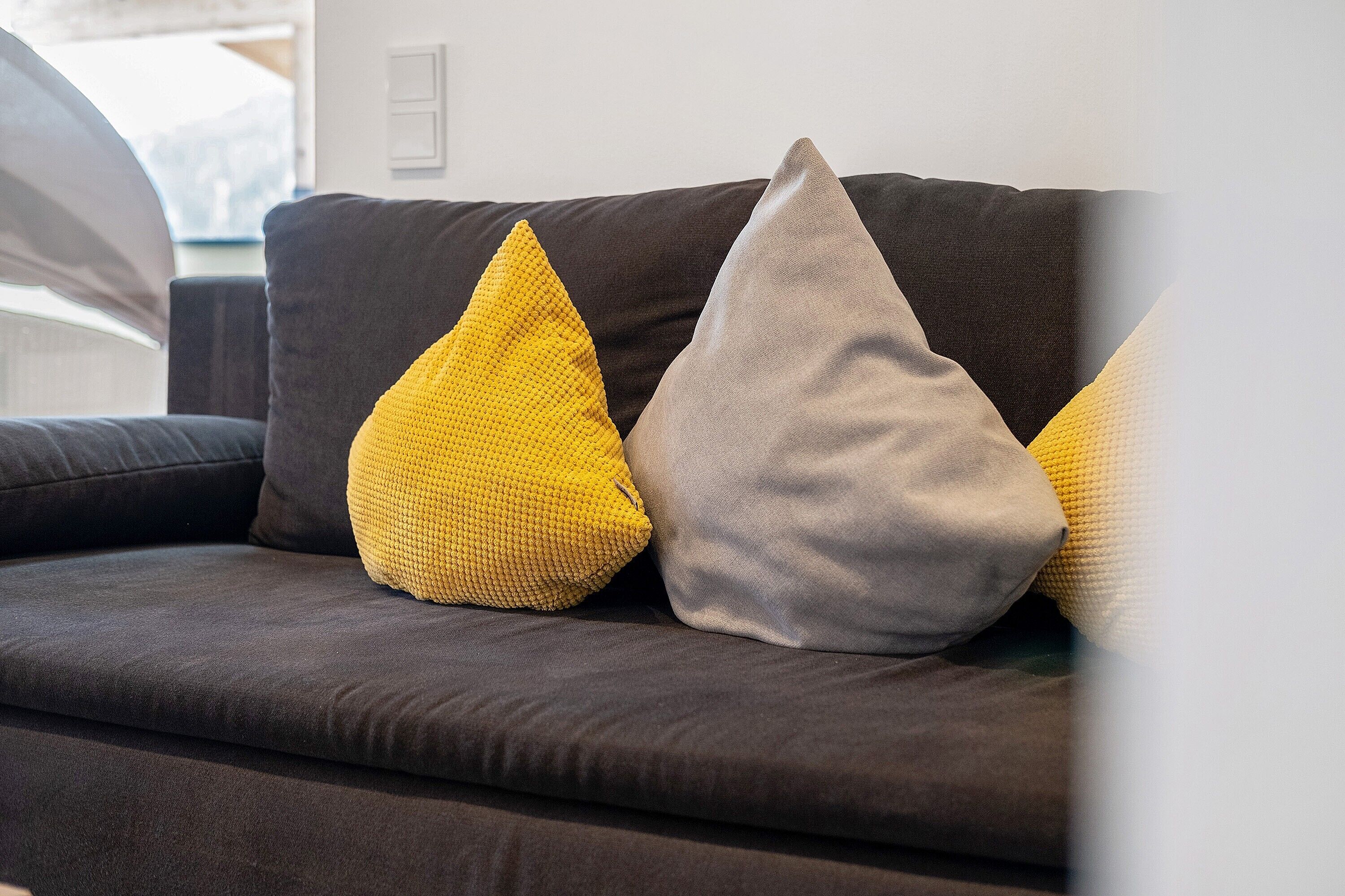 A dark-colored sofa with three cushions, one yellow and two gray, in a room with a white wall and a window.