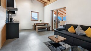 Ein modernes Interieur mit Küche, Essbereich und Wohnbereich, mit Holzbalkendecke, einer Couch mit Kissen und Blick durch eine Glastür.