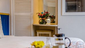 A neatly made bed with a floral bedspread, a bedside table with a coffee maker, and a vase with flowers on a shelf.