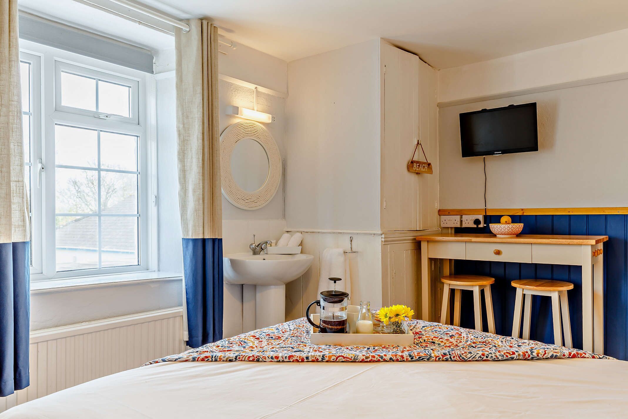 A bedroom with a bed, a bedside table with a tray of breakfast items, a sink, and a TV mounted on the wall.