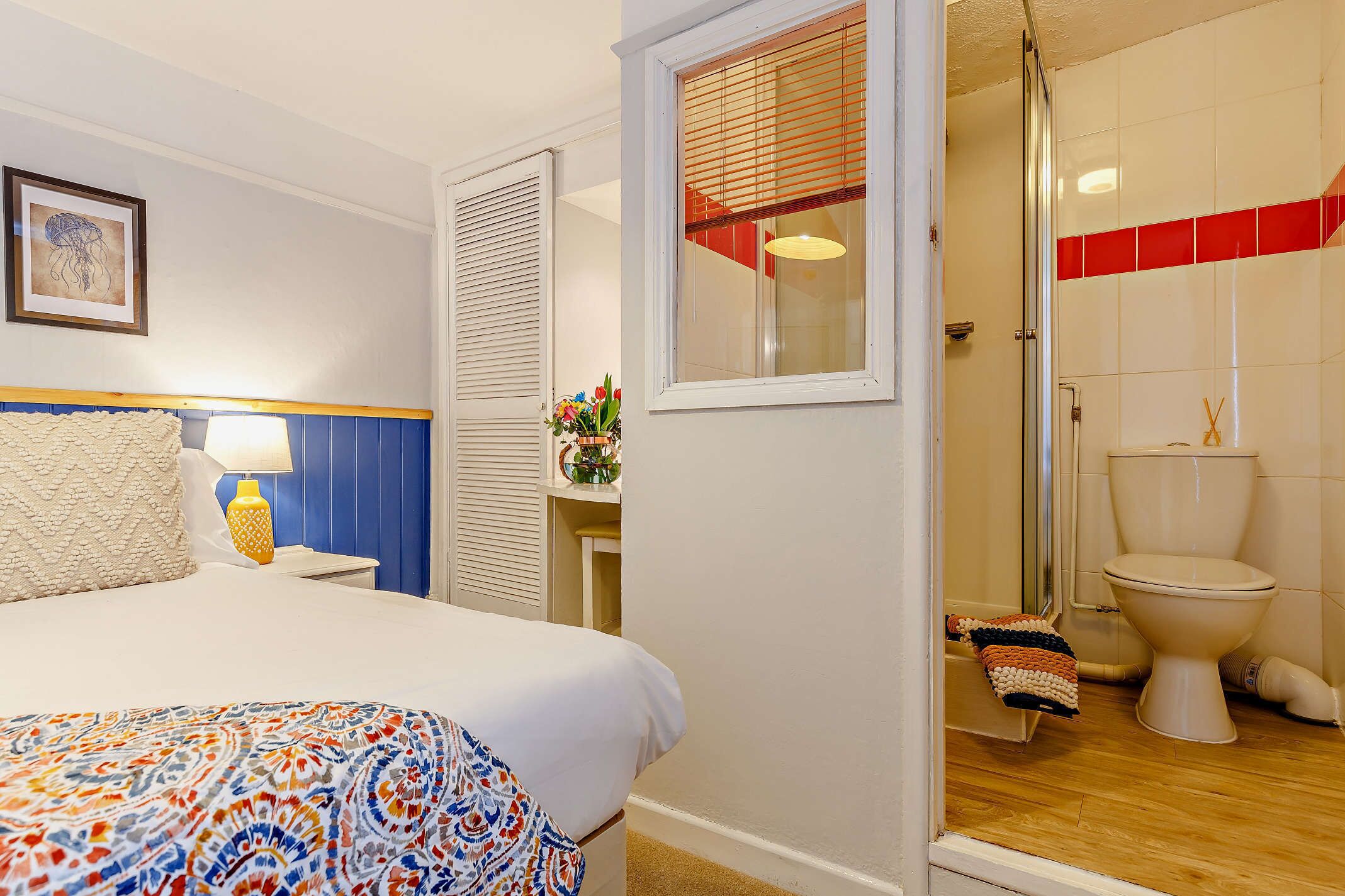 A hotel room with a bed, a bedside table with a lamp, a window with blinds, a bathroom with a toilet, and a shower.