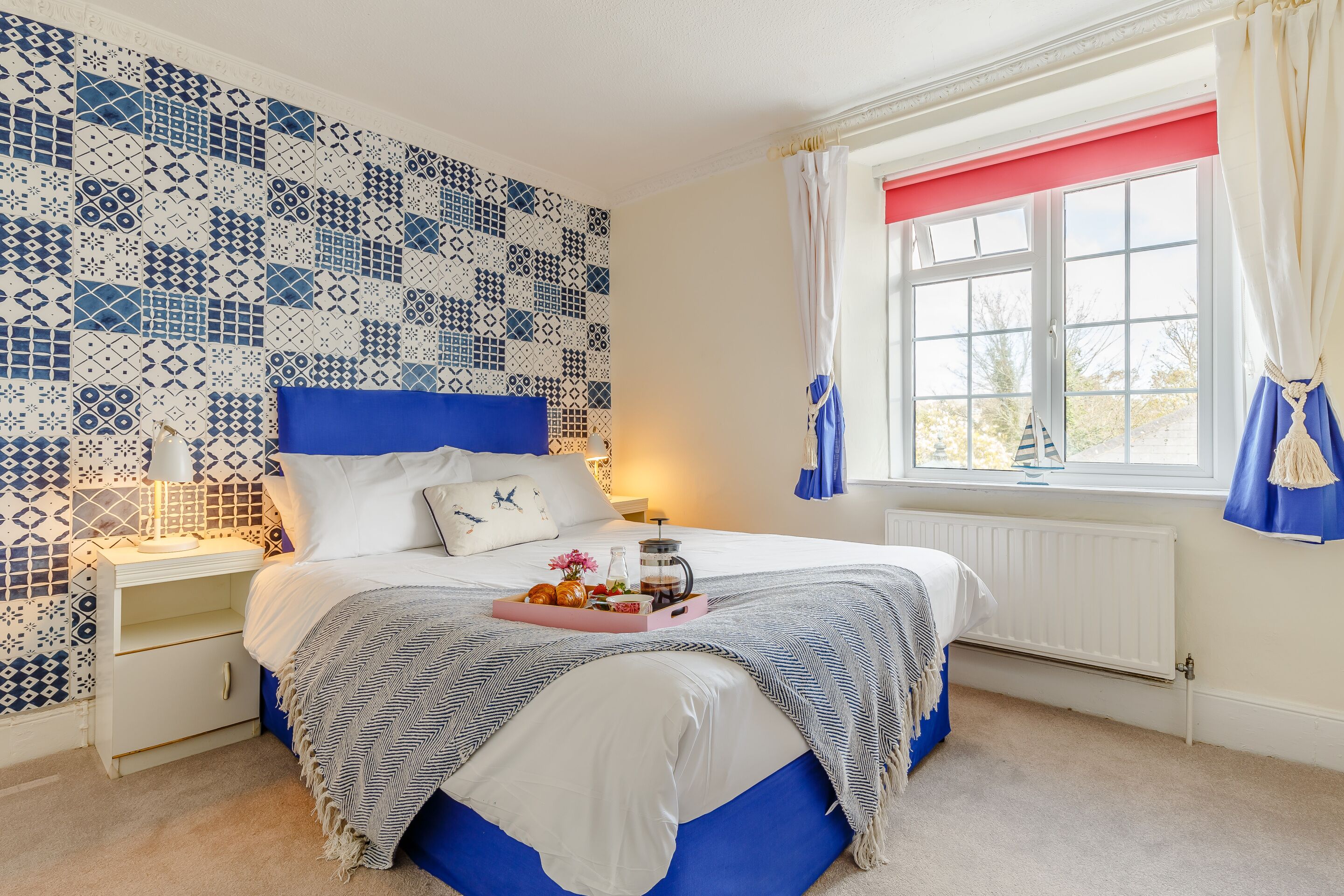 A bedroom with a patterned blue and white wall, a bed with a blue headboard, a bedside table with a lamp, and a window with curtains.