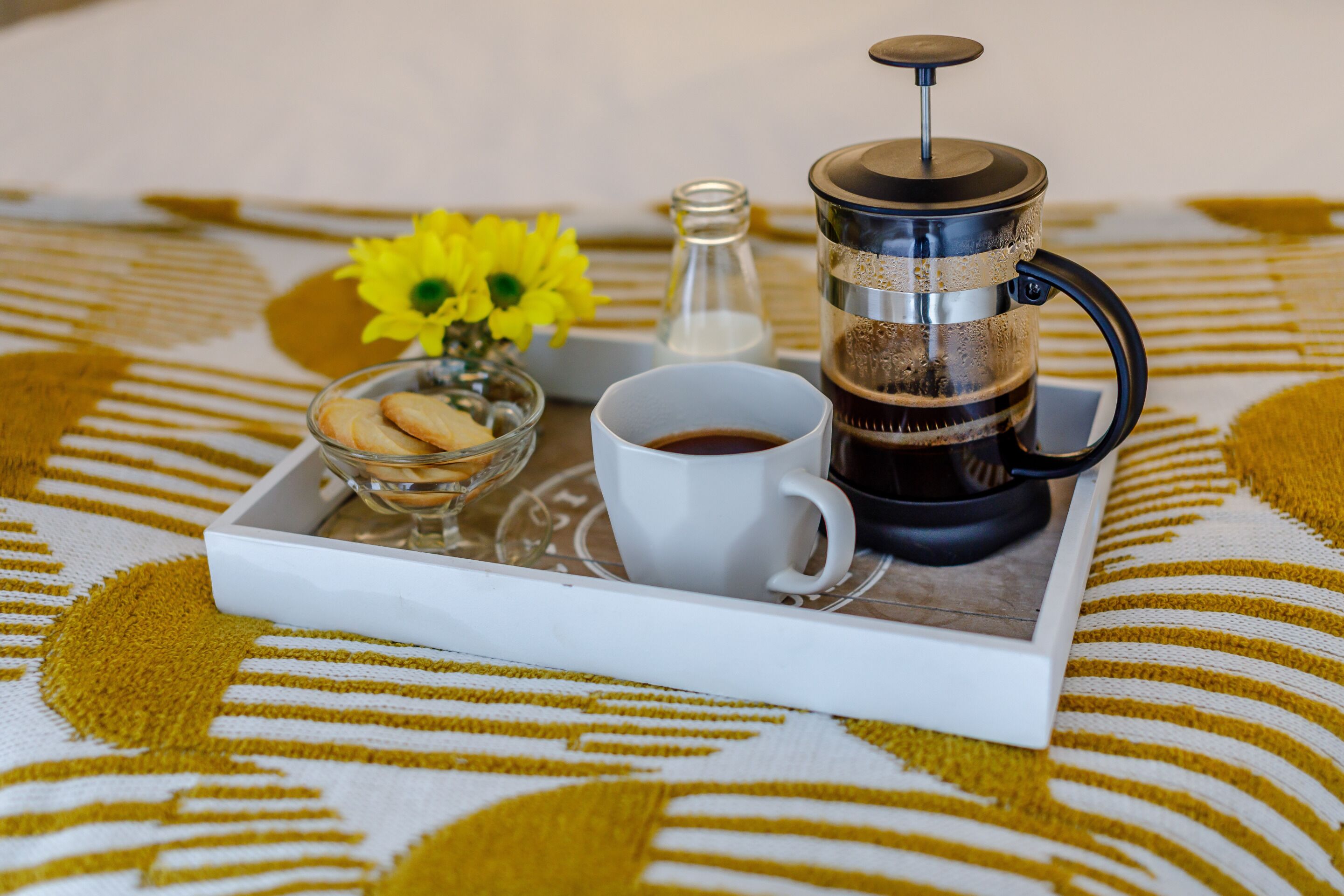 Ein Tablett mit einer Kaffeemaschine, einer Kaffeetasse, einem Glas Wasser, einer kleinen Schale Kekse und einer Vase mit gelben Blumen auf einer gemusterten Tischdecke.