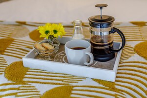 Una bandeja con cafetera, una taza de café, un vaso de agua, un pequeño cuenco de galletas y un florero con flores amarillas sobre un mantel estampado.