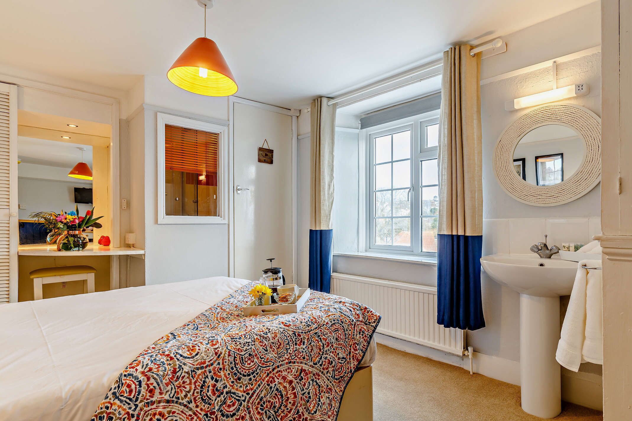 Habitación de hotel con cama, mesita de noche, lámpara, ventana con cortinas y baño con lavabo y espejo.
