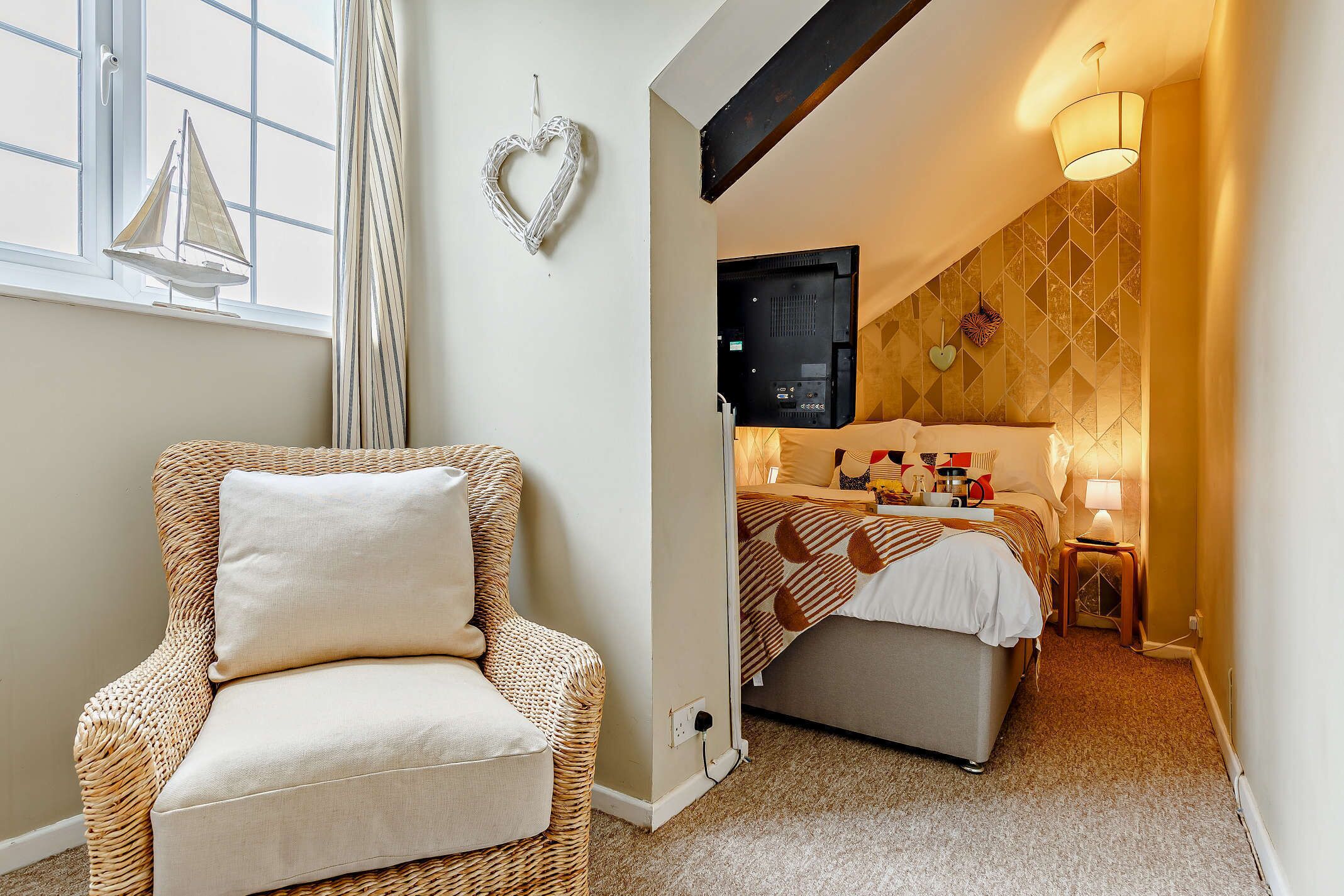 Una habitación acogedora con una silla de mimbre, una cama con una manta estampada, un televisor montado en la pared y una decoración con forma de corazón.