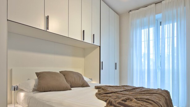 Un dormitorio con cama, armarios blancos y una ventana con cortinas traslúcidas.