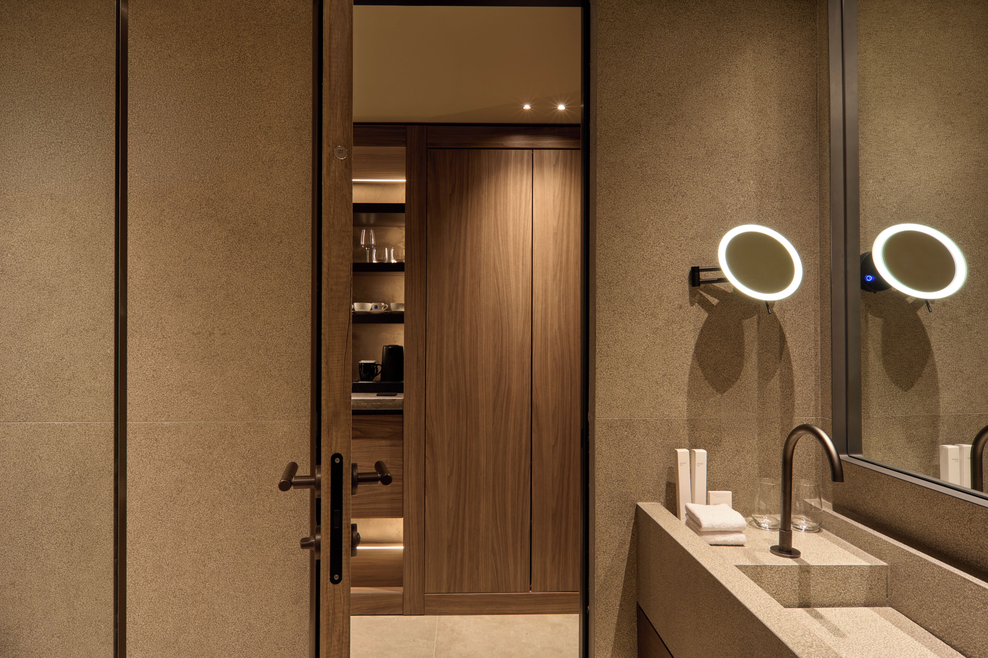 A modern bathroom with a vanity, a mirror with a light, and a wooden door leading to a closet.