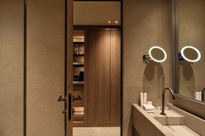 A modern bathroom with a vanity, a mirror with a light, and a wooden door leading to a closet. - Nur Coreon Signature Hotel (Athens)
