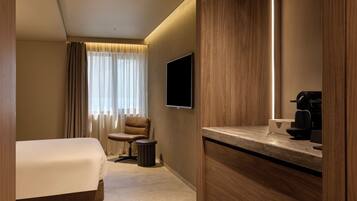 Quarto de hotel com cama, uma cadeira, escrivaninha com cafeteira, televisão e janela com cortinas.
