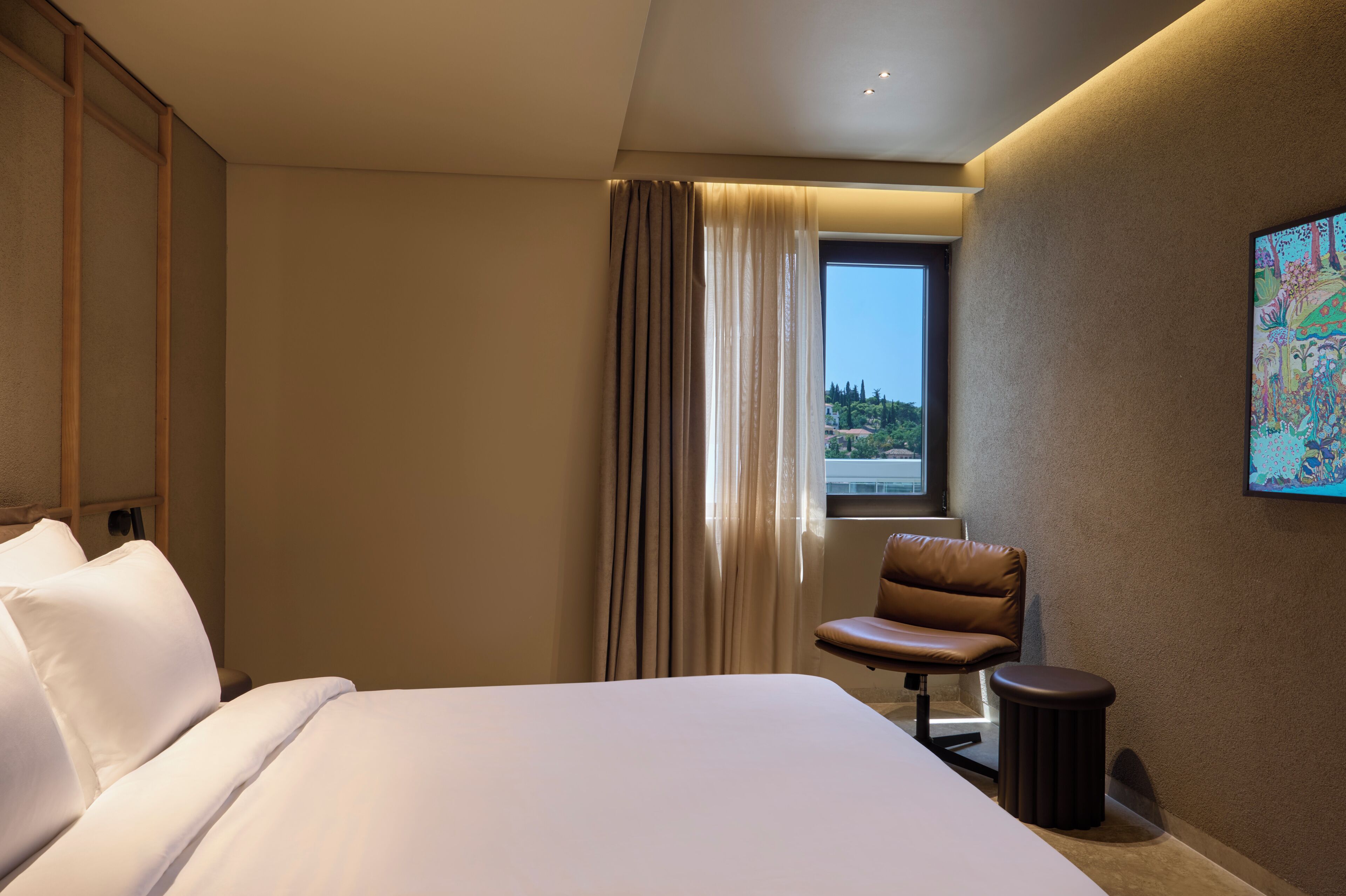 Ein Hotelzimmer mit Bett, einem Sessel, einem kleinen Tisch, einem Fenster mit Vorhängen und einem Gemälde an der Wand.