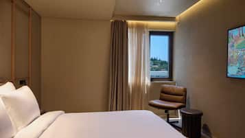 A hotel room with a bed, a chair, a small table, a window with curtains, and a painting on the wall.