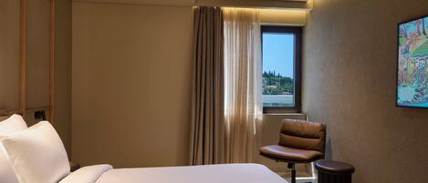 A hotel room with a bed, a chair, a small table, a window with curtains, and a painting on the wall.