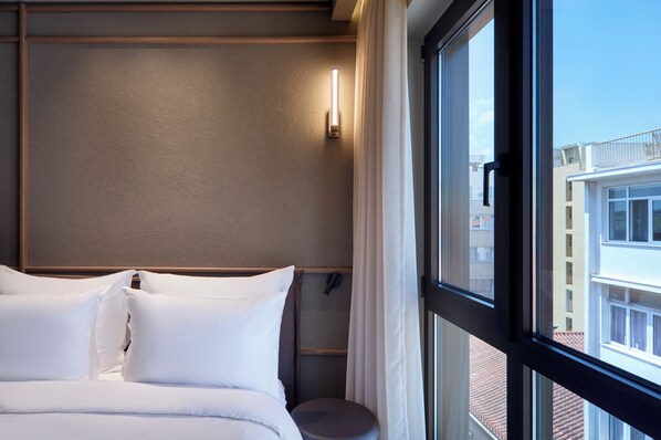 Una camera d'albergo con un letto, un comodino, una finestra con tende e vista sugli edifici.