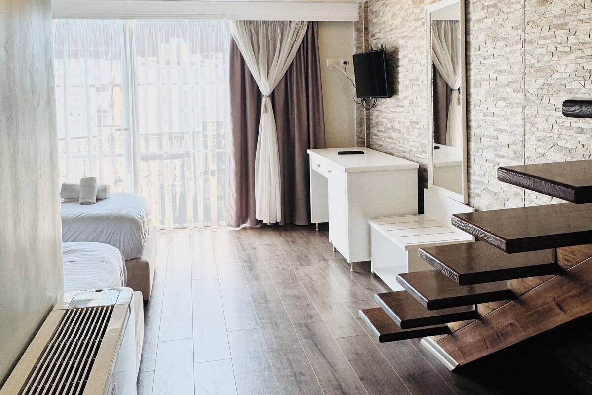 A modern hotel room with a wooden ceiling, a large window with curtains, a bed, a desk, and a mirror.