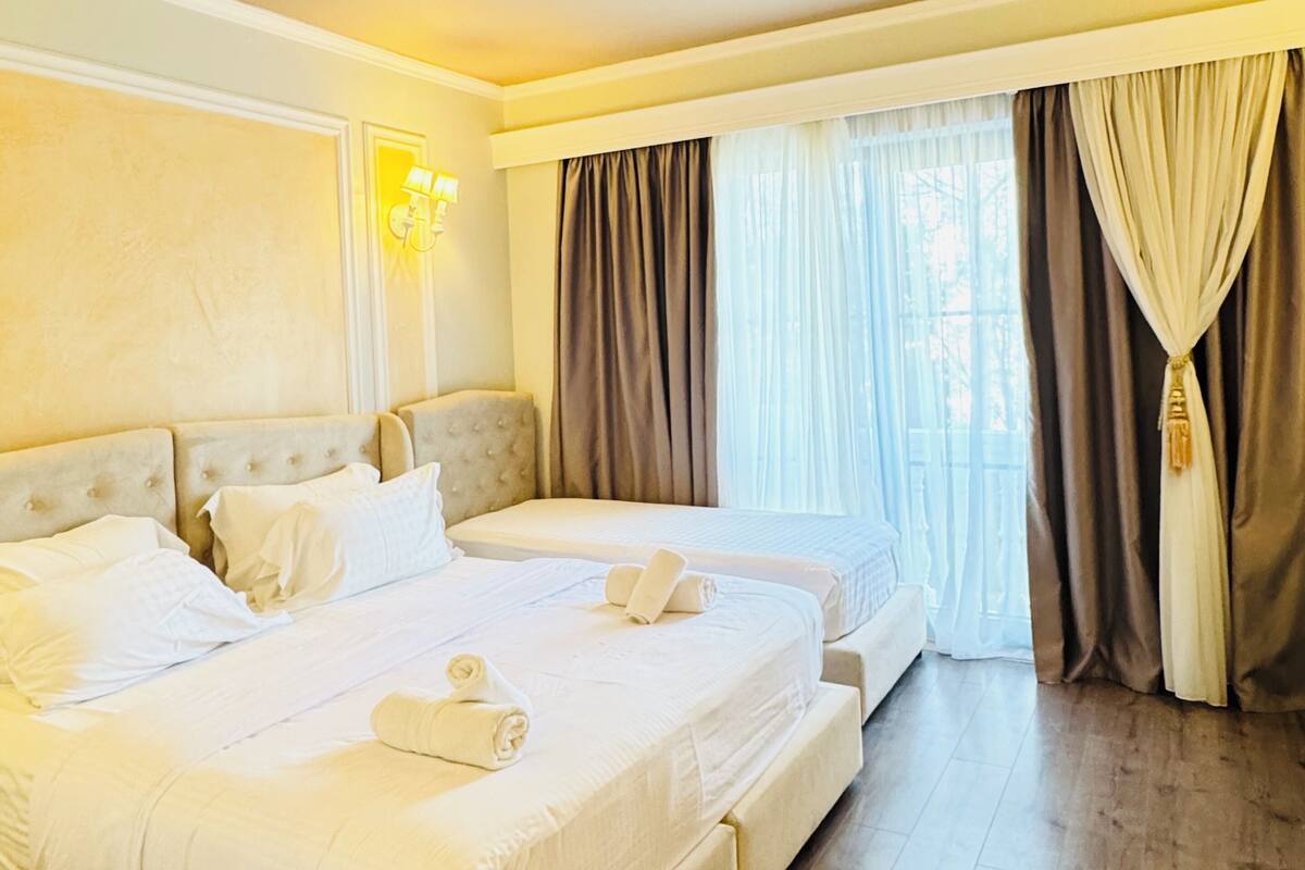 A hotel room with two beds, a large window with curtains, a wall-mounted lamp, and a wooden floor.