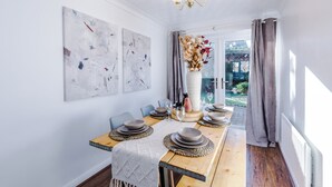 A dining area with a long wooden table set for four, two wall-mounted abstract paintings, and a chandelier.
