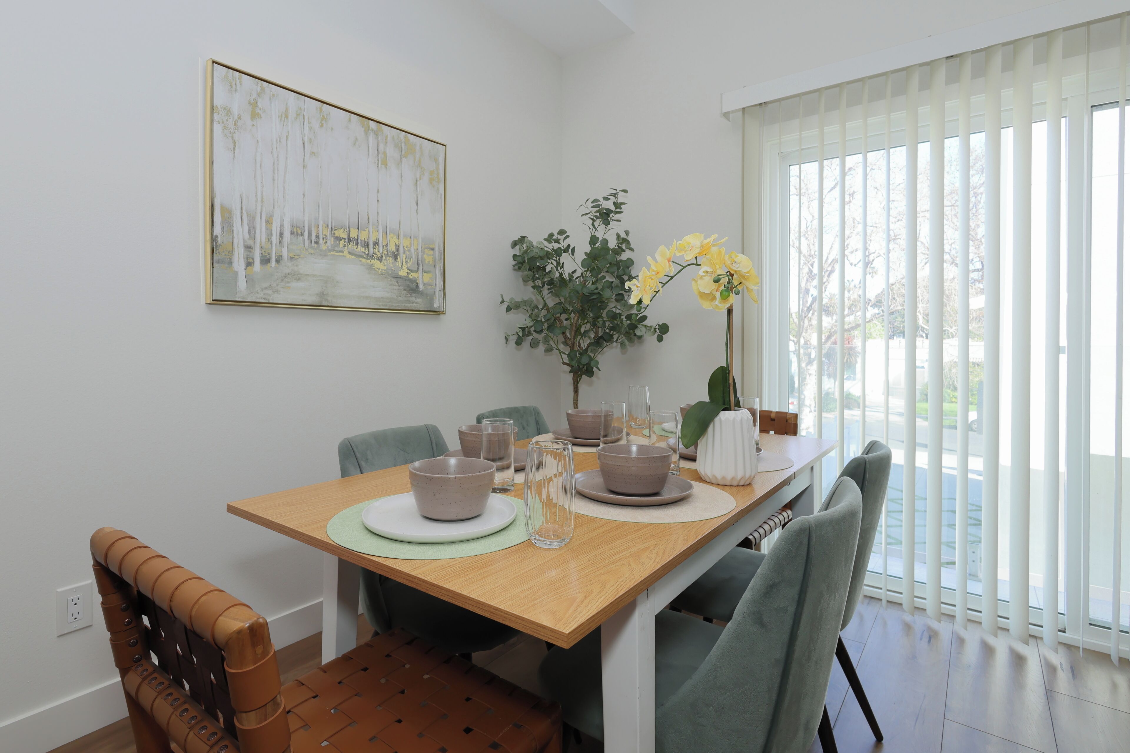 A dining area with a wooden table, chairs, and a vase of yellow flowers. There is a painting on the wall and a window with blinds.