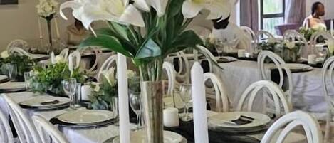 A dining area with white chairs, a table set with plates, glasses, and a centerpiece of white flowers.