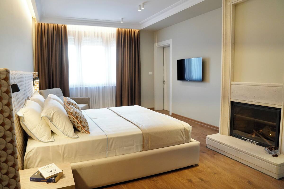 Ein modernes Hotelzimmer mit einem großen Bett, einem Flachbildfernseher, einem Kamin und Blick auf die Stadt durch große Fenster.