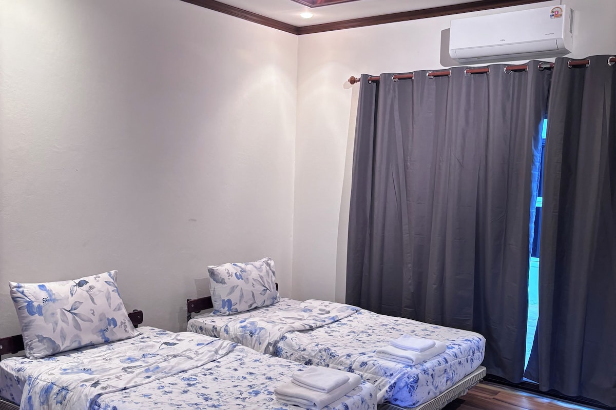 A room with two beds, a ceiling fan, an air conditioning unit, and a window with curtains.