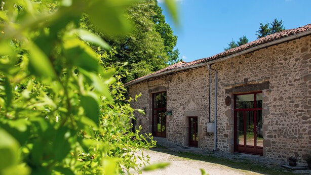 A stone building with a tiled roof, a gravel path, and a green bush in the foreground.