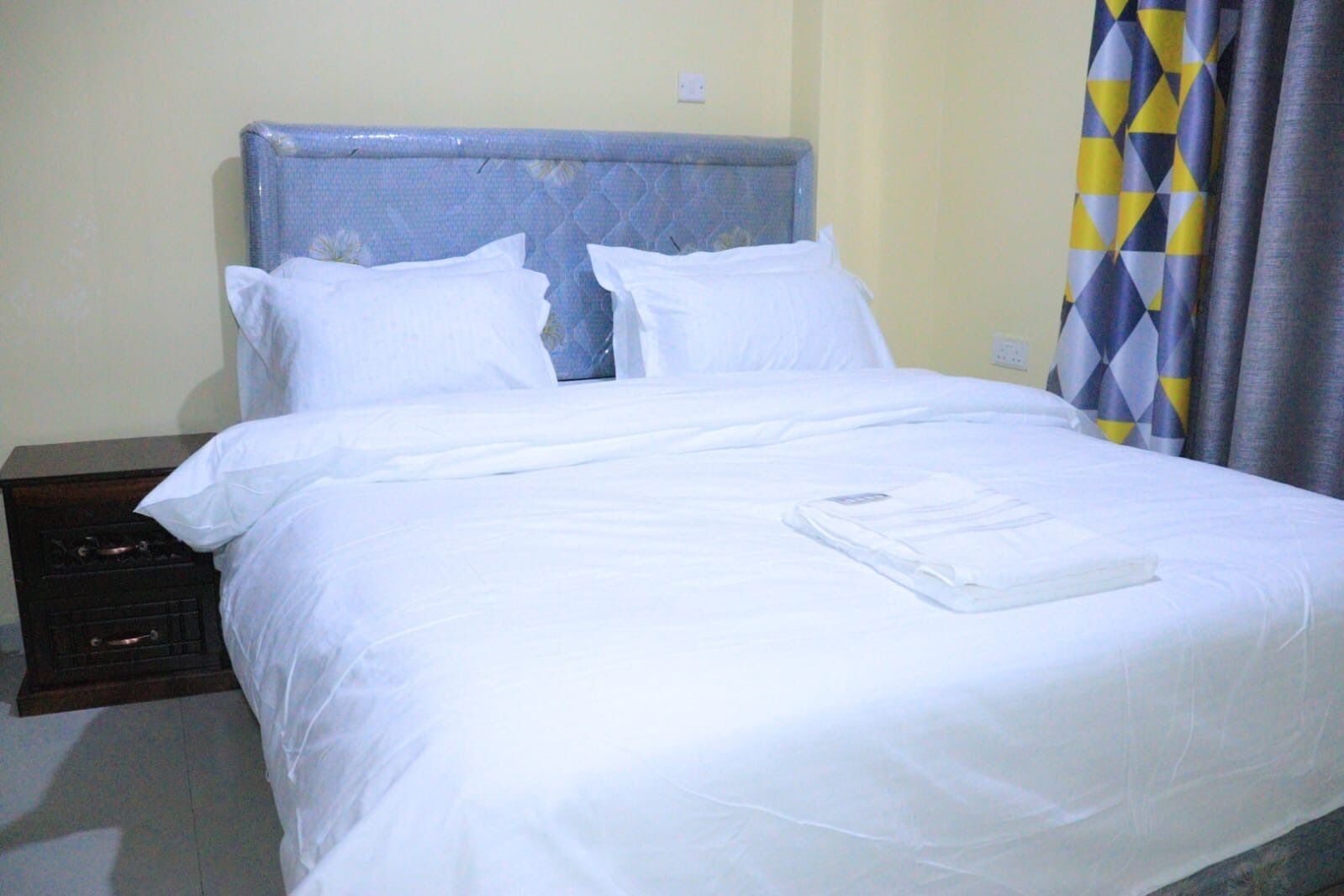 A neatly made bed with white linens, a wooden nightstand to the left, and a patterned curtain to the right.