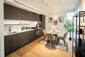 A modern kitchen with dark wood cabinets, a white countertop, and a dining area with a round table and chairs. - Elegant 2BR London Escape • A/C • Gym Access (London)
