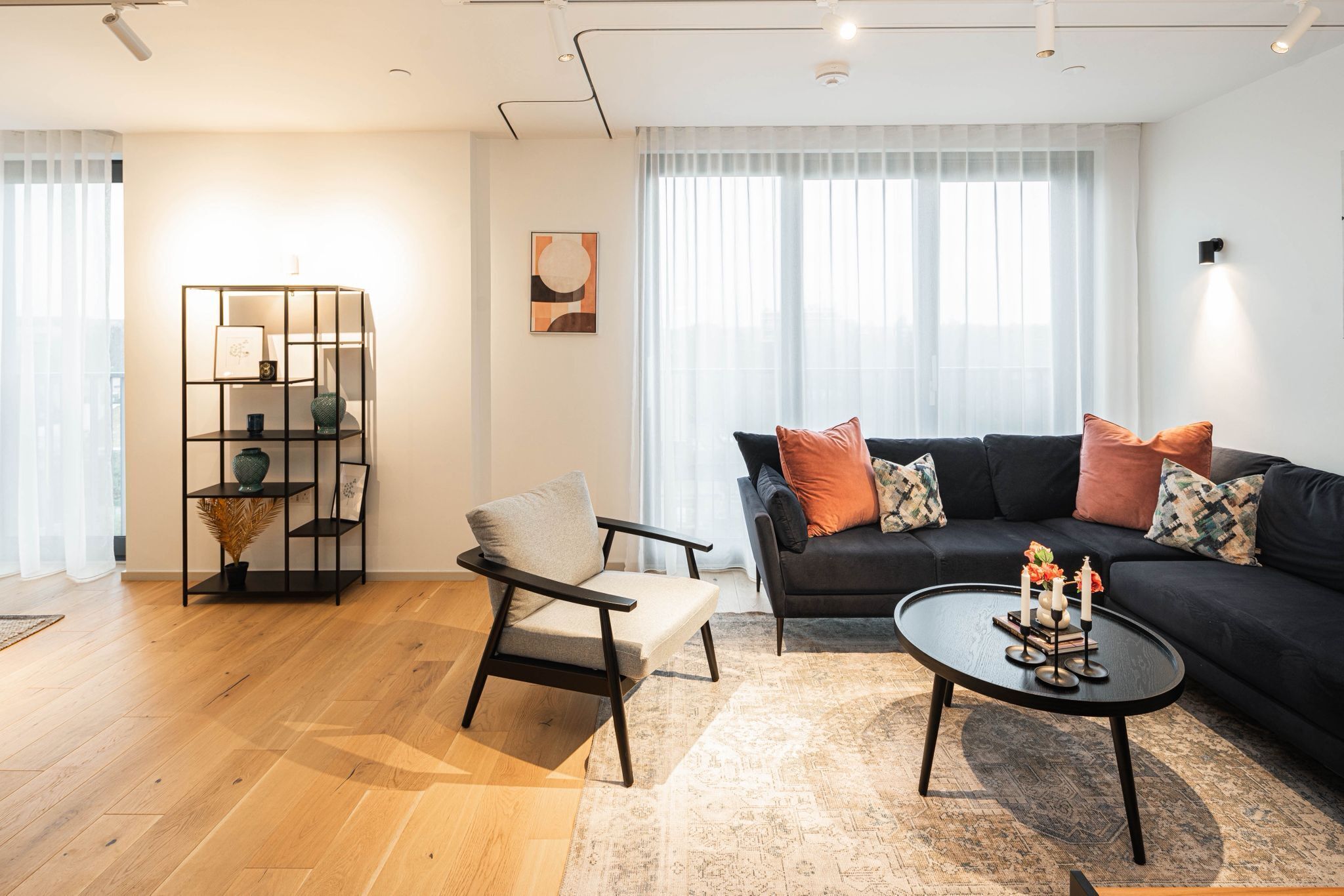 A modern living room with a black sofa, a grey armchair, and a coffee table. There is a black bookshelf with decorative items and a framed picture on the wall.
