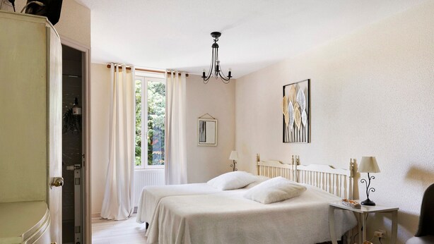 Un dormitorio con dos camas, una araña, una mesita de noche con lámpara, una silla y una ventana con cortinas.