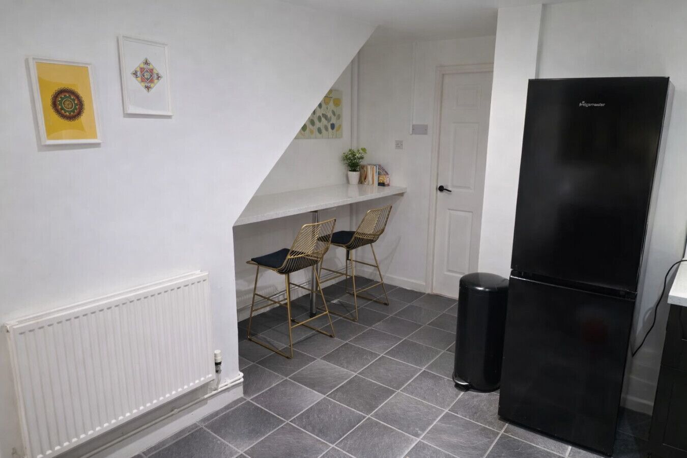 A compact kitchen with a black refrigerator, two chairs, a white radiator, and a small shelf with items.